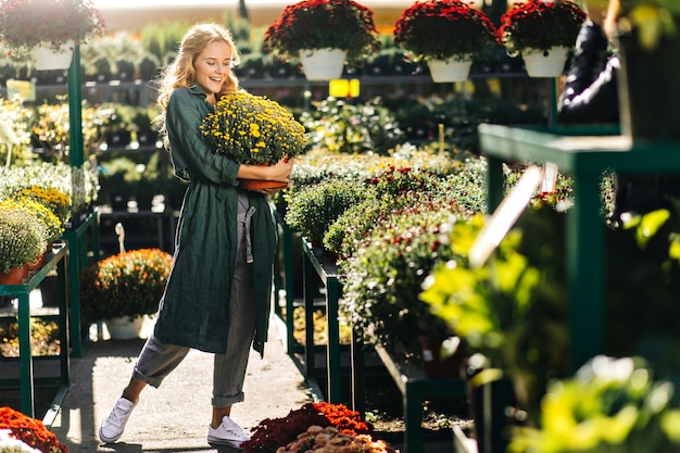 Young woman with beautiful blond hair and gentle smile, dressed in green robe with belt is working in greenhouse