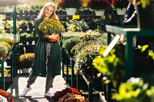 Young woman with beautiful blond hair and gentle smile, dressed in green robe with belt is working in greenhouse