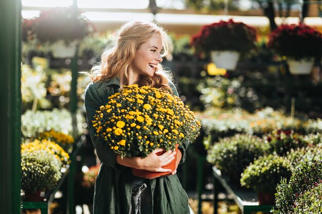 Young woman with beautiful blond hair and gentle smile, dressed in green robe with belt is working in greenhouse