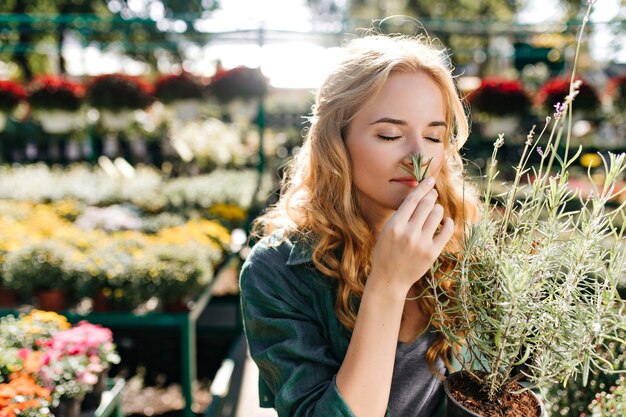 Young woman with beautiful blond hair and gentle smile, dressed in green robe with belt is working in greenhouse