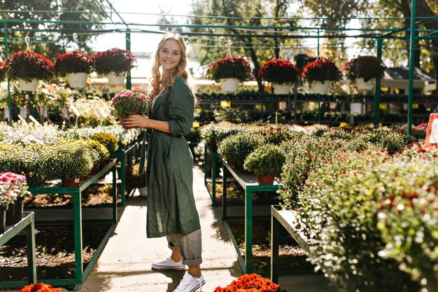 Young woman with beautiful blond hair and gentle smile, dressed in green robe with belt is working in greenhouse