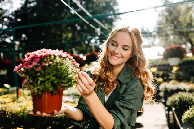 Young woman with beautiful blond hair and gentle smile, dressed in green robe with belt is working in greenhouse