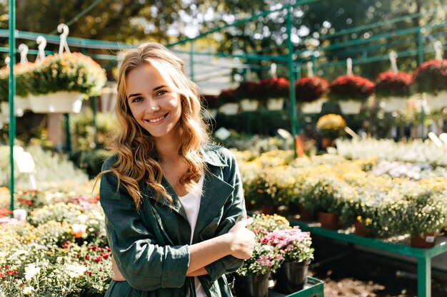 Young woman with beautiful blond hair and gentle smile, dressed in green robe with belt is working in greenhouse