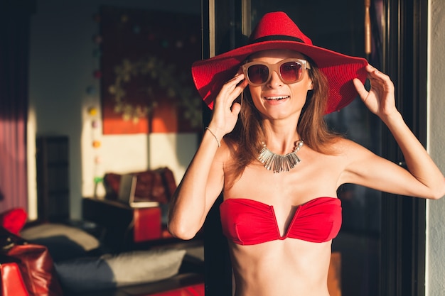 Young woman with beautiful slim body wearing red bikini swimsuit, straw hat and sunglasses relaxing on tropical villa resort during vacation in Asia, skinny figure, summer style trend accessories