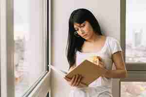 Free photo young woman with beverage reading near windows