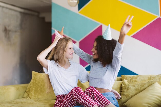 Young woman with blond hair and pretty african american woman with dark curly hair in birthday caps joyfully looking at each other while spending time together with colorful wall on background