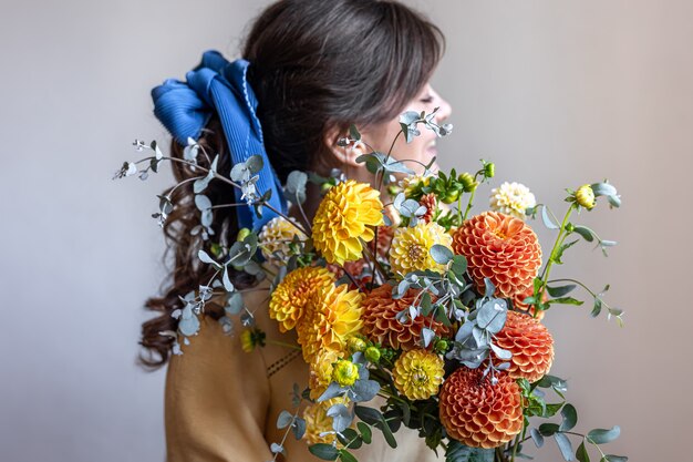 彼女の髪に青いリボンを持つ若い女性、黄色とオレンジ色の菊の花束、灰色の背景を保持しています。