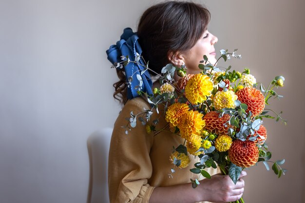 Young woman with a blue ribbon in her hair, holding a bouquet of yellow and orange chrysanthemums, gray background.