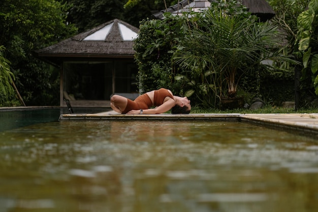 Young woman with body positive appearance practicing yoga alone on deck by the pool in tropical island of Bali, Indonesia. Sport, fitness, healthy lifestyle concept.