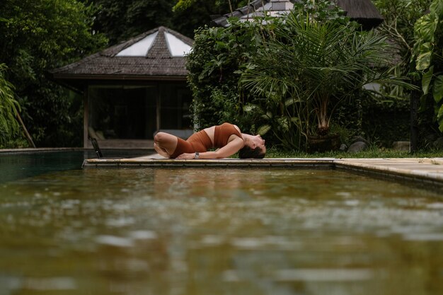 인도네시아 발리의 열대 섬 수영장 옆 갑판에서 혼자 요가를 하는 긍정적인 몸을 가진 젊은 여성. 스포츠, 피트니스, 건강한 라이프스타일 컨셉입니다.