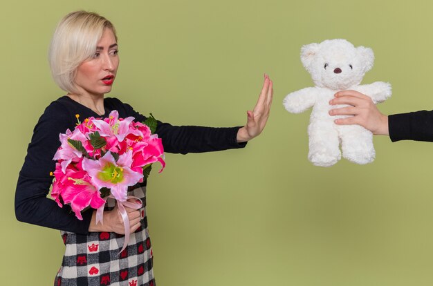 young woman with bouquet of flowers looking confused making stop gesture while receiving teddy bear as a gift