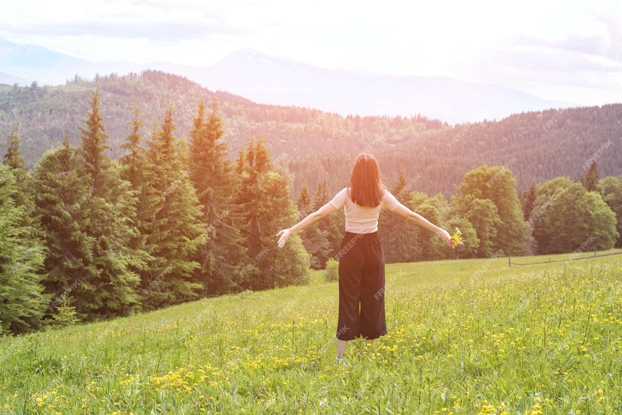 Premium Photo | Young woman with a bouquet of flowers and wide open arms standing in the meadow. forest and mountains in the background. back view