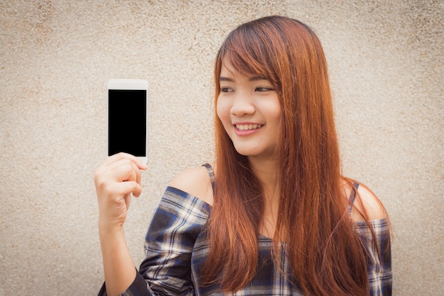 Young woman with brown hair smiling showing a blank smartphone screen standing on concrete wall background. Vintage effect style pictures.