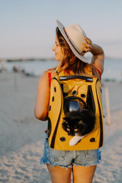 海岸のバックパックに猫を抱く若い女性。ペットと一緒に旅行のコンセプト。舷窓付きのバックパックの猫。