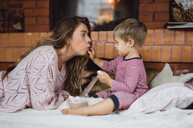 young woman with a child by the fireplace. mom and son drink cocoa with marshmallows 