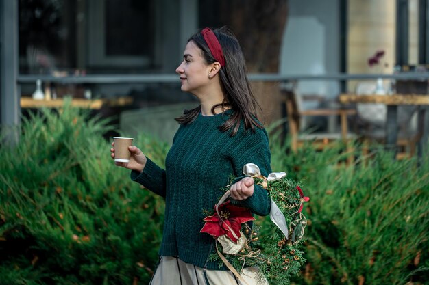 Молодая женщина с рождественским венком и чашкой кофе на прогулке по городу
