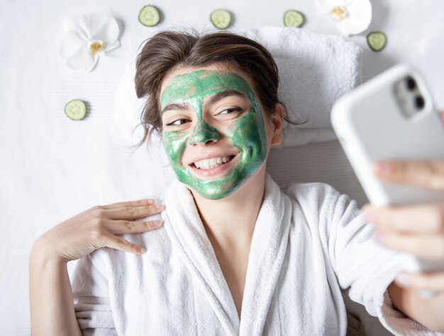 Young woman with a cosmetic mask on her face makes a selfie on a smartphone.