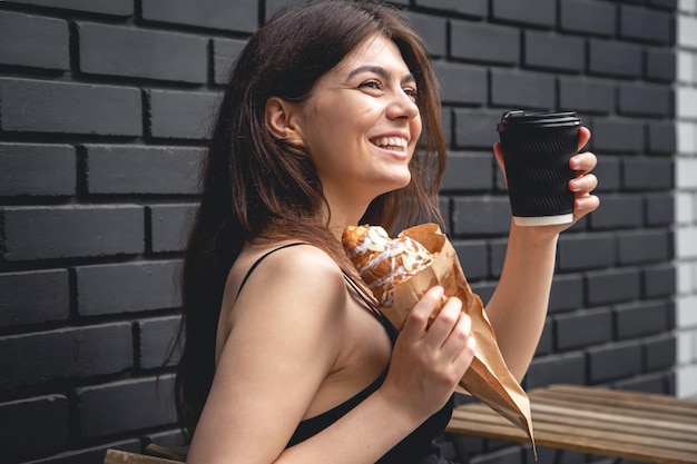 Una giovane donna con un croissant e una tazza di caffè contro un muro di mattoni neri