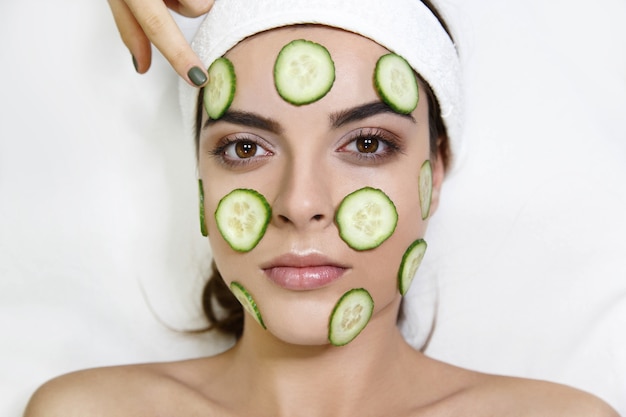 Young woman with cucumber mask lies on white table in spa saloon