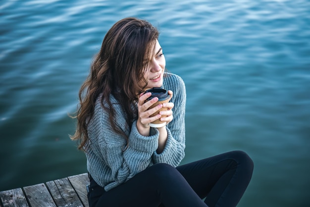 Free photo a young woman with a cup of coffee sits by the river