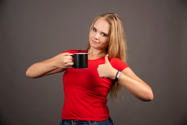 Young woman with cup showing thumbs up on black wall.