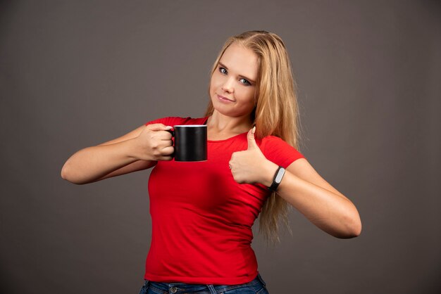 Young woman with cup showing thumbs up on black wall.