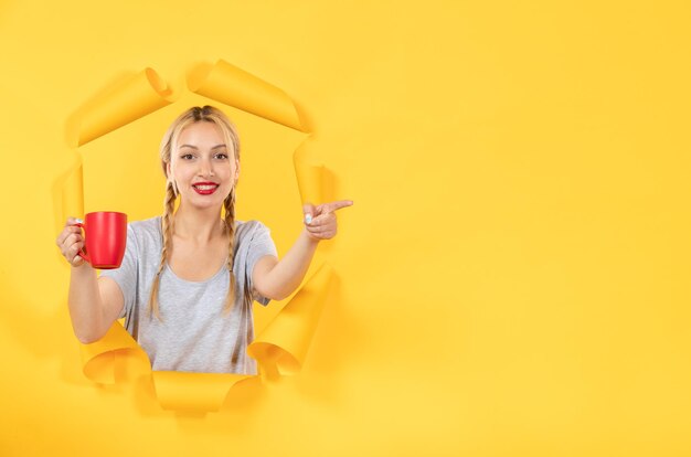 Young woman with cup of tea pointing at something on yellow background advertising