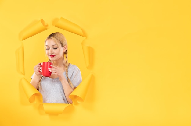 Young woman with cup of tea on torn yellow paper background advertising shopping