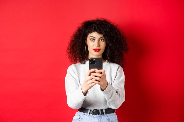 Young woman with curly hair, recording video on smartphone, taking photo on mobile phone and looking at camera, standing on red background