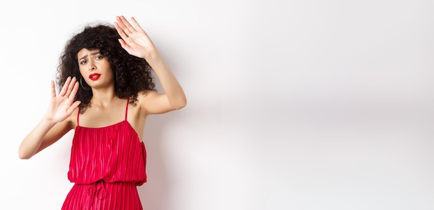 Young woman with curly hair and red dress asking to stop block someone raising hands defensive prote