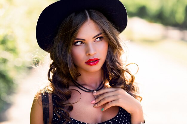 young woman with dark curly hair in stylish elegant black hat posing in the park