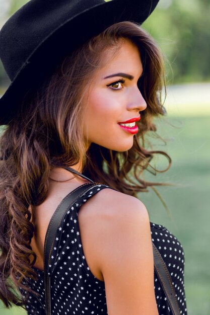 young woman with dark curly hair in stylish elegant black hat posing in the park