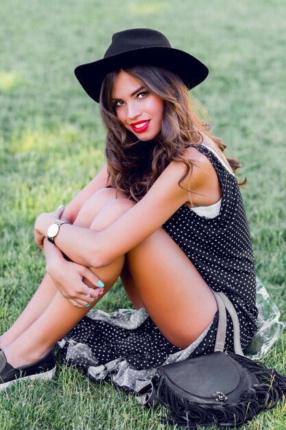 young woman with dark curly hair in stylish elegant black hat posing in the park