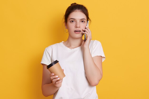 Young woman with dark hair wearing white casual t shirt, posing against yellow wall