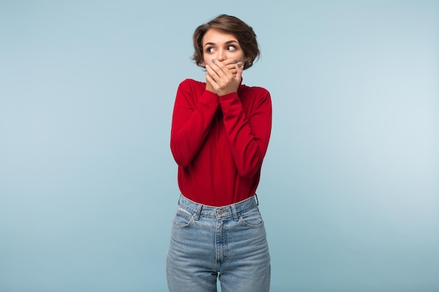 Young woman with dark short hair in red sweater and jeans covering mouth with hands while thoughtfully looking aside over blue background