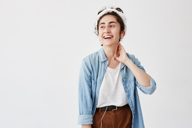 Young woman with dark and wavy hairstyle, wears denim shirt, looks happily aside, laughs, has good mood, listens to audio book with earphones, isolated on white wall