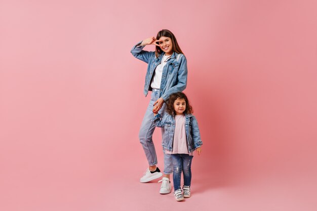 ピースサインを示す娘と若い女性。ピンクの背景に子供と手をつないで素晴らしいスタイリッシュな女性のスタジオショット。