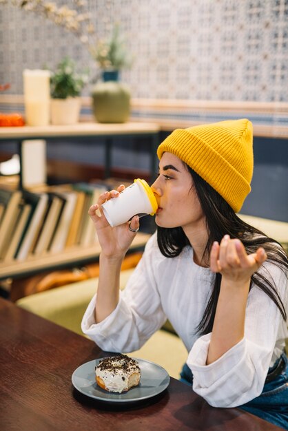 カフェのテーブルでカップから飲むプレートのデザートを持つ若い女