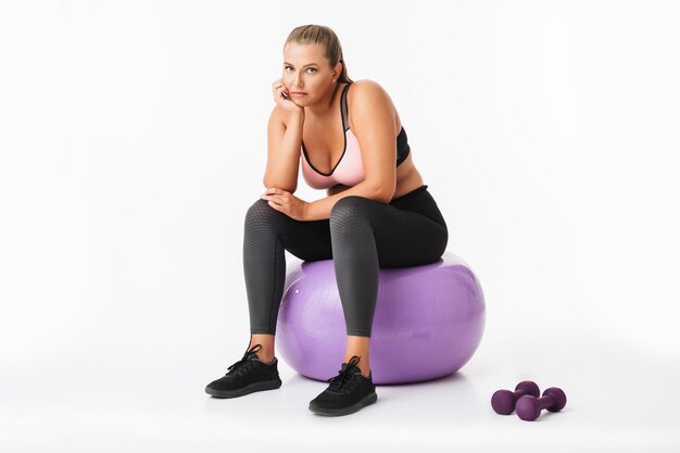 Молодая женщина с избыточным весом в спортивном топе и леггинсах сидит на фитнес-мяче с гантелями рядом, задумчиво глядя в камеру на белом фоне изолированы