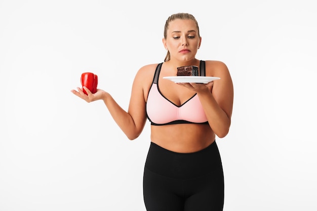 Foto gratuita giovane donna con peso in eccesso nella parte superiore sportiva scegliendo premurosamente tra pepe e torta al cioccolato su sfondo bianco isolato