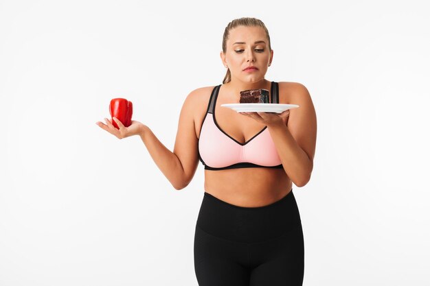 Young woman with excess weight in sporty top thoughtfully choosing between pepper and chocolate cake over white background isolated