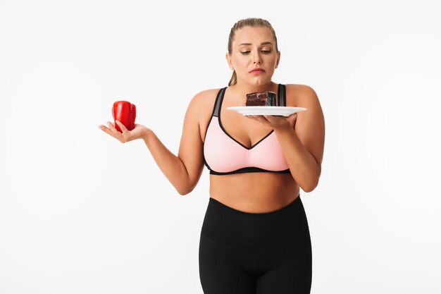 Молодая женщина с избыточным весом в спортивном топе задумчиво выбирает между перцем и шоколадным тортом на белом фоне изолированы
