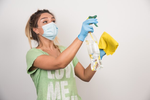 Young woman with facemask and supplies spraying on white wall. 