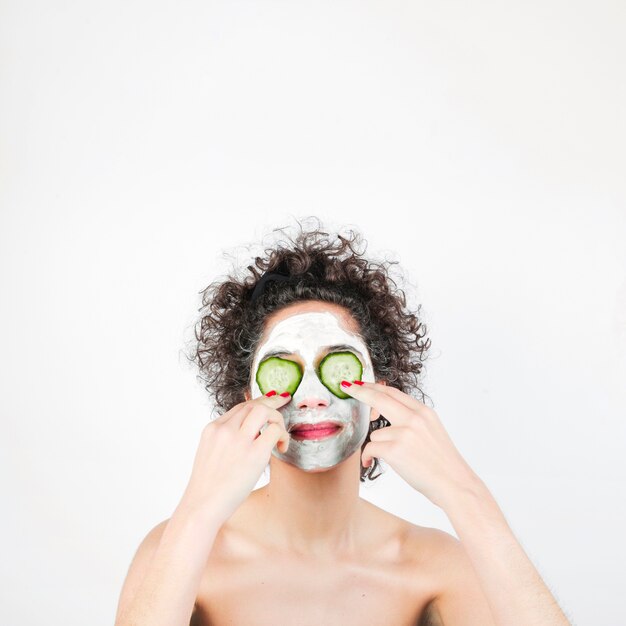 Young woman with facial mask and cucumber slices on face against white background