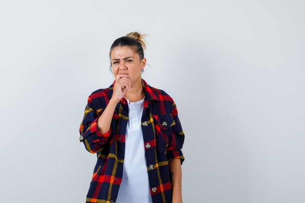 Young woman with finger in mouth in checkered shirt and looking puzzled , front view.