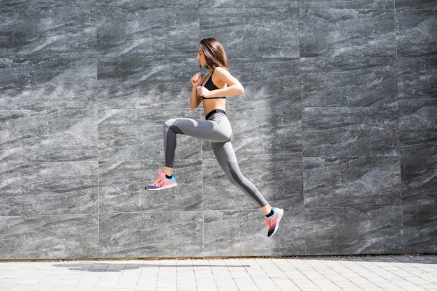 灰色の壁にジャンプして走っているフィットボディの若い女性。