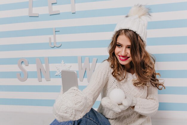 Young woman with gentle smile and blue eyes poses for selfie, showing snowball in her hand. Indoor portrait of girl in knitted white clothes