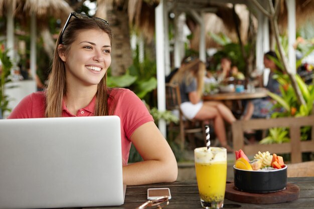 Young woman with glasses on her head smiling joyfully, resting at cafe and browsing internet using laptop computer, sitting at table with fruit shake and cell phone, people having lunch