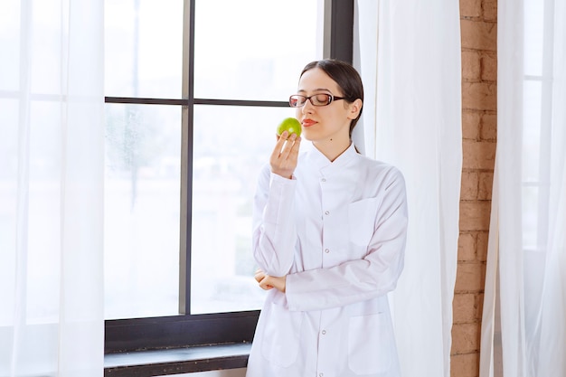 창 근처 녹색 사과 먹는 실험실 코트에 안경을 쓴 젊은 여자.