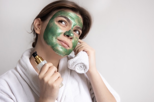 A young woman with a green beauty mask on her face holds a towel and a care product, copy space.